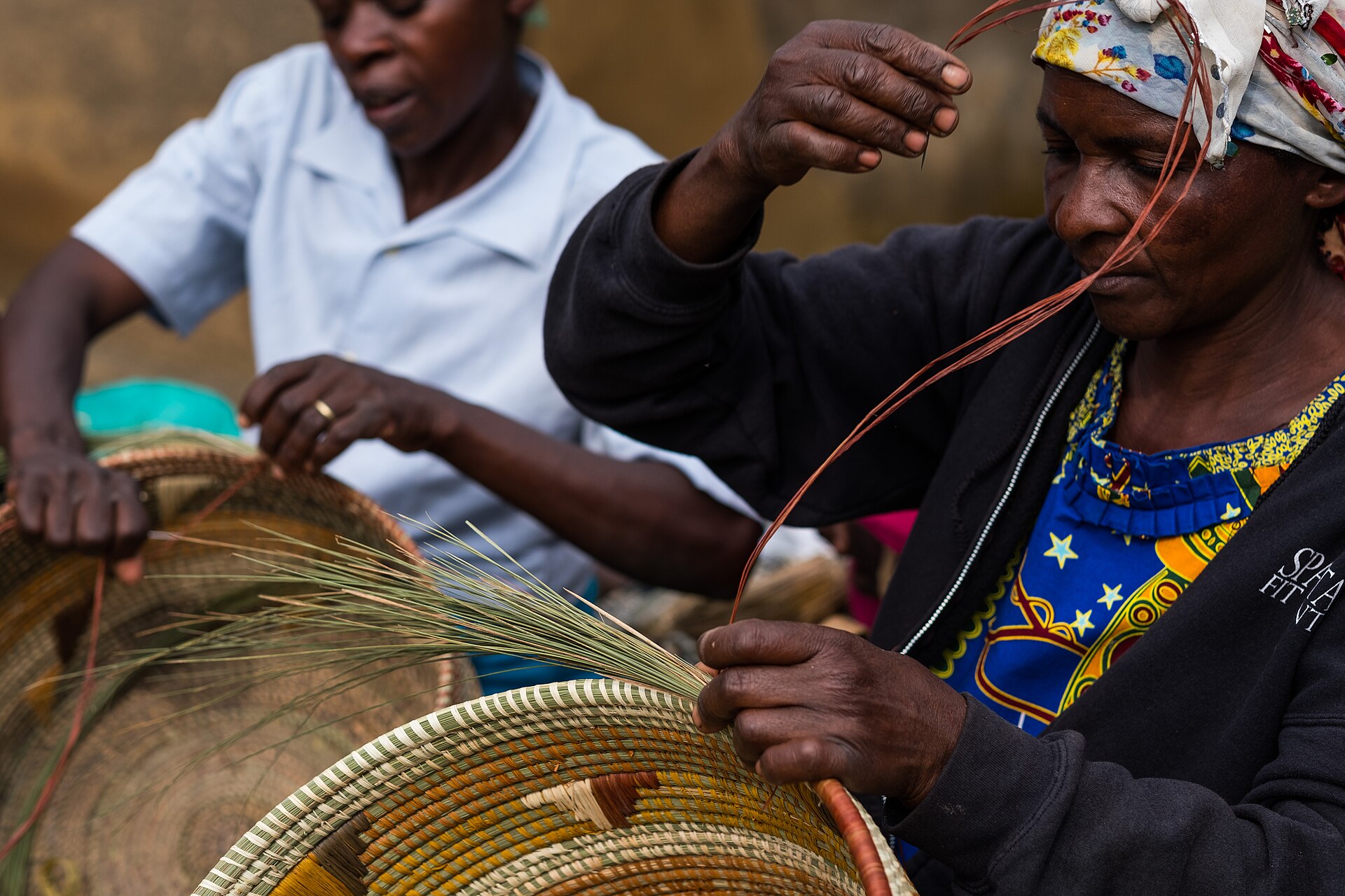 Two women weaving baskets side by side, an older woman in the foreground and another behind her.