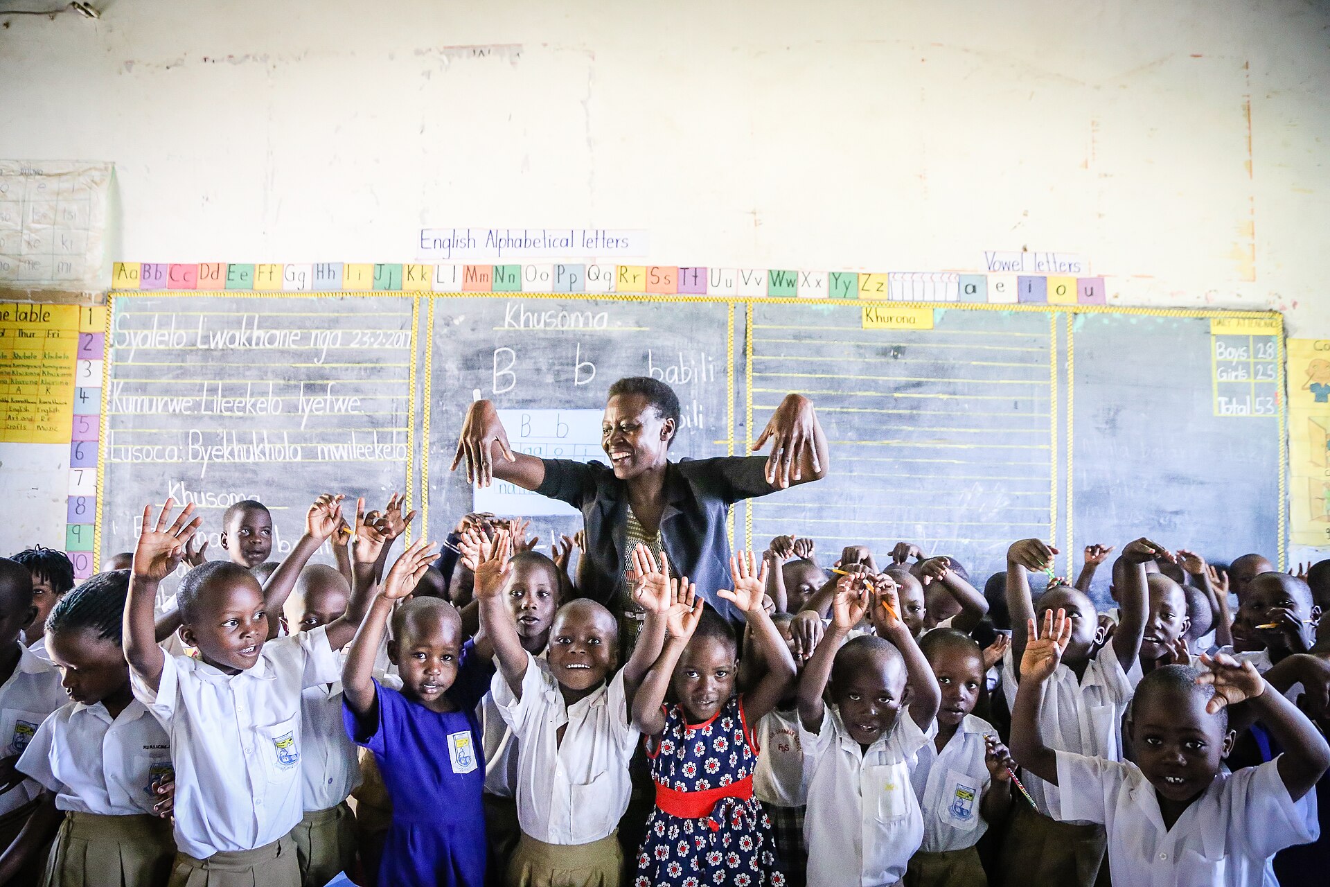 A teacher in front of a chalkboard, surrounded by smiling primary school pupils with their hands raised.
