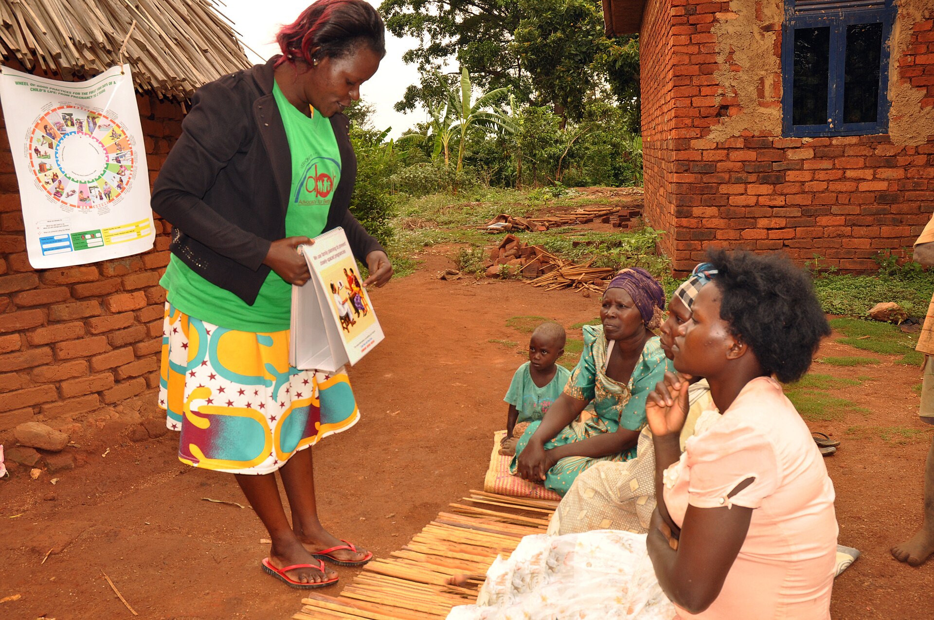 A community health worker holding a flip chart and talking with a family seated on a mat outside their brick home.